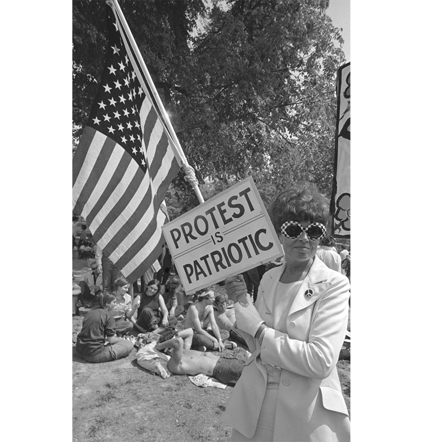 A photograph from the book The Activist's Media Handbook, David Fenton. A woman holds a sign that reads "Protest is Patriotic" and the U.S Flag.